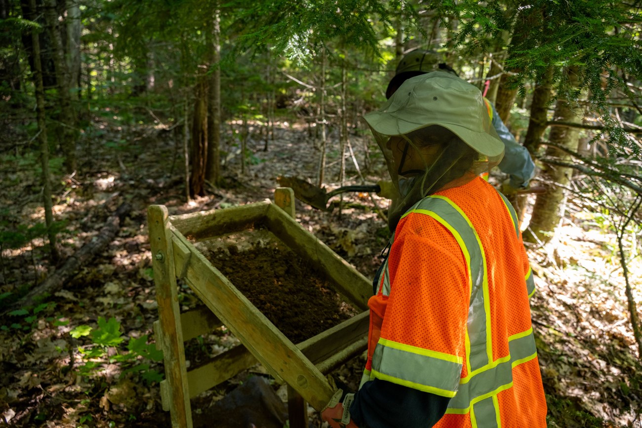 An archeologist wearing a hi-vis orange work vest and a bug net over her head while using a soil sifter.