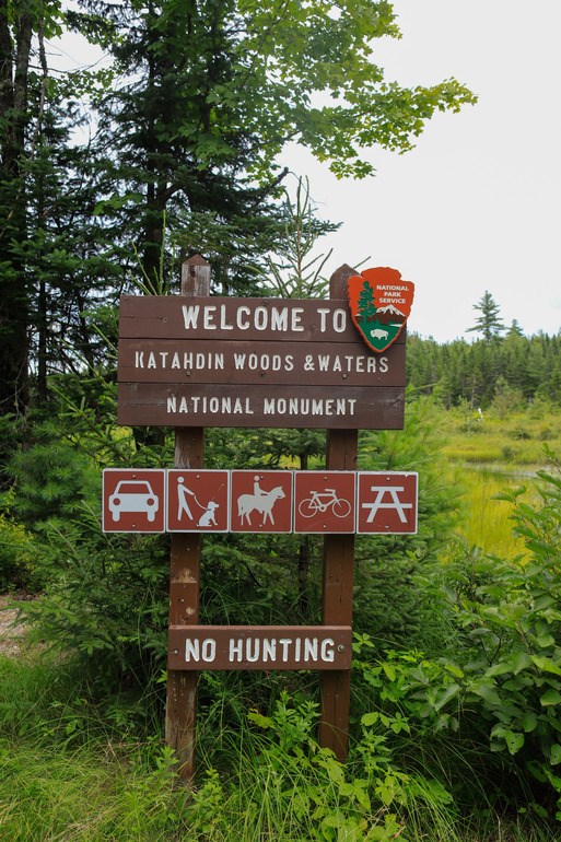 A brown wooden welcome sign with the NPS Arrowhead reads "Welcome to Katahdin Woods & Waters National Monument"