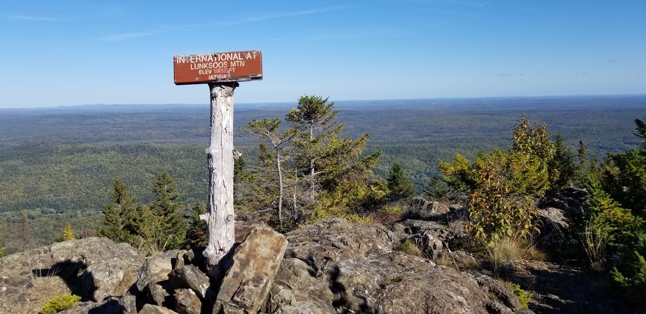 a pile of rocks sits at the summit of a mountain. In the center of the image, a sign reading "International AT, Lunksoos Mountain" is propped in the rock pile. An expanse of green trees and blue skies fill the background.