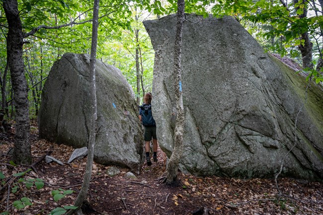 Backpacker hikes between narrow gap in two large rock formations, surrounded by green trees