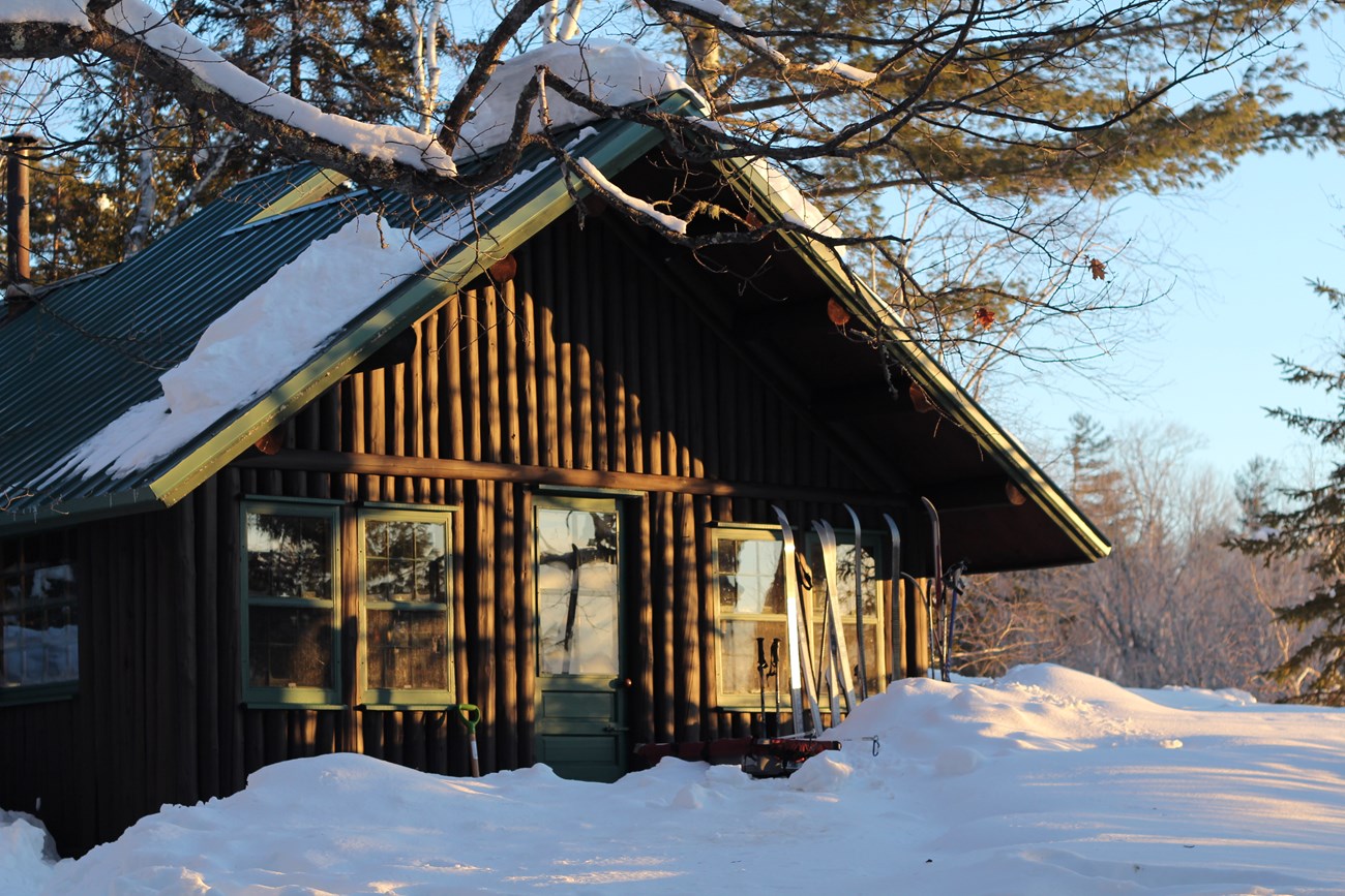 A small wooden cabin on a blue sky day surrounded by thick snow. The sun glares agains the windows with pairs of cross country skis standing outside.