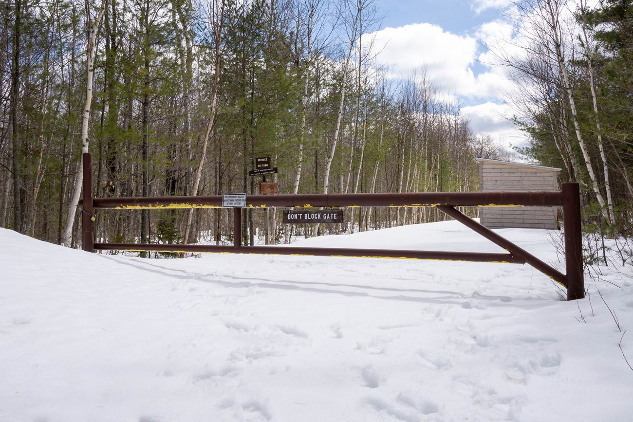 A large brown metal gate is closed and locked over a snowy road in the woods. It is a blue sky day with large white patchy clouds.