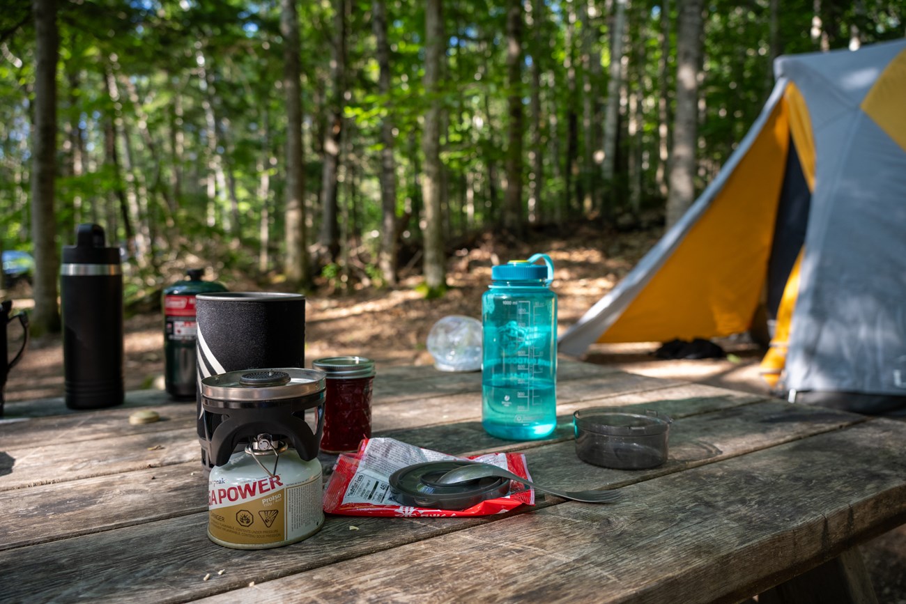 A picnic table with food items and cooking materials on it sits at a summer campsite with a yellow tent in the background. Green trees can be seen in the distance.