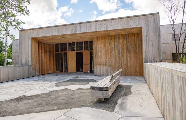 East-facing entrance of Tekαkαpimək Contact Station. The right door of the rectangular wooden entrance is propped open on a sunny day with patchy clouds. A wooden bench oriented perpendicular to building faces south.