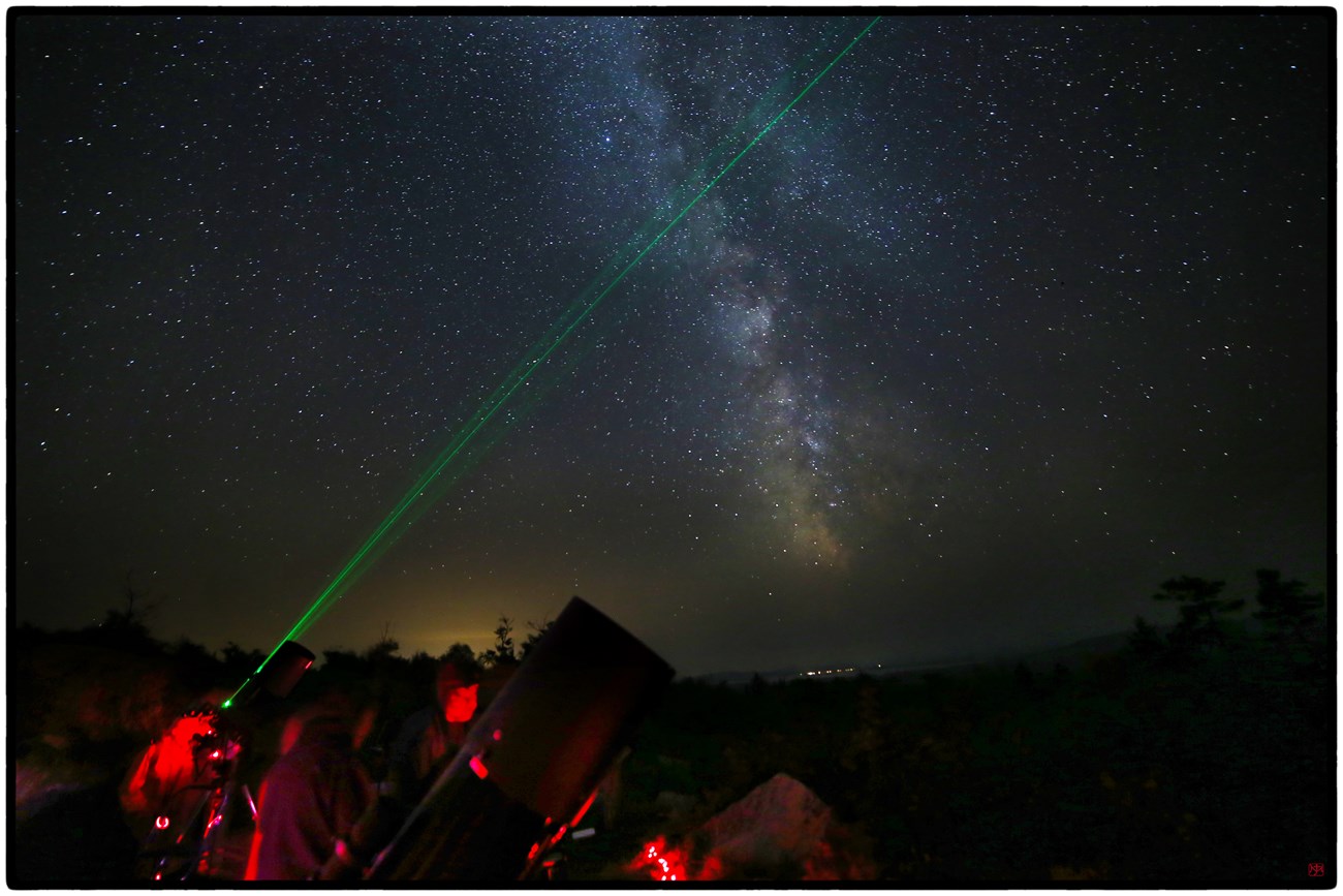 View of the Milky Way from the Loop Road Overlook at Stars Over Katahdin 2017. In the foreground a large telescope is visible. A green laser points to the stars and people's faces reflect the red lights used to preserve night sky vision.