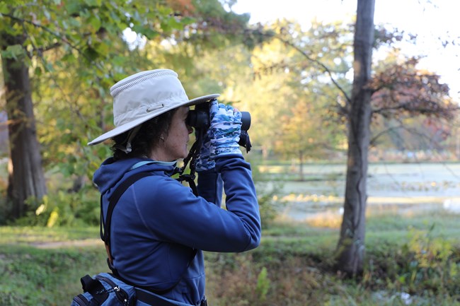 A woman, viewed from the side, wears a light-colored, wide-brimmed hat, a blue hooded jacket, and patterned gloves. She is holding binoculars up to her eyes, looking towards a body of water in the middle ground that reflects the sky.