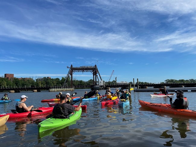 Kayakers are floating in the Anacostia River, there is a train bridge behind them.