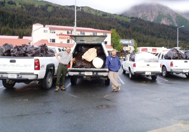 Two park tech stand next to the rear of two pick-up trucks filled with garbage bags.