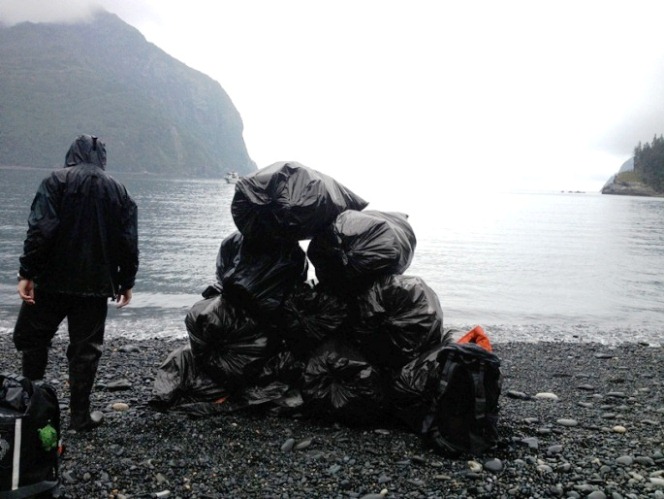 A park tech stands on a beach, next to a pyramid of ten garbage bags. 