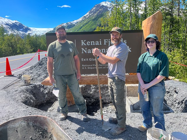 Three people including a youth staff pose with the Kenai Fjords National Park sign under construction