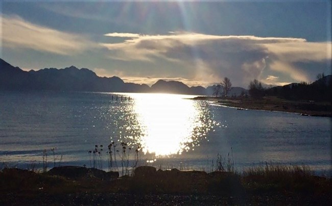 Sun shining on the water.  Clouds rise above mountains in the distance.