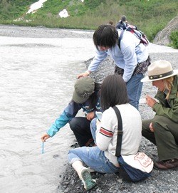 Three students from Obhiro, Japan, stand on the edge of Exit Creek, measuring the temperature of  the water and following the directions of an Education Ranger.