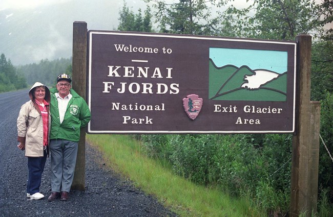 An older couple stands next to an Exit Glacier welcome sign, in the early 1990s.