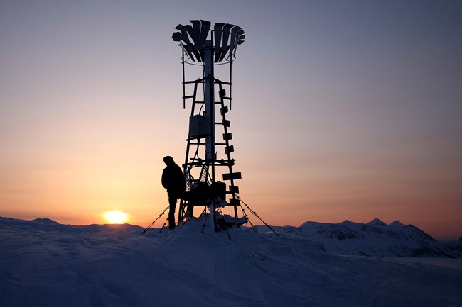 A researcher stands next to a tall weather station on a snow covered field with the sun setting in the background.