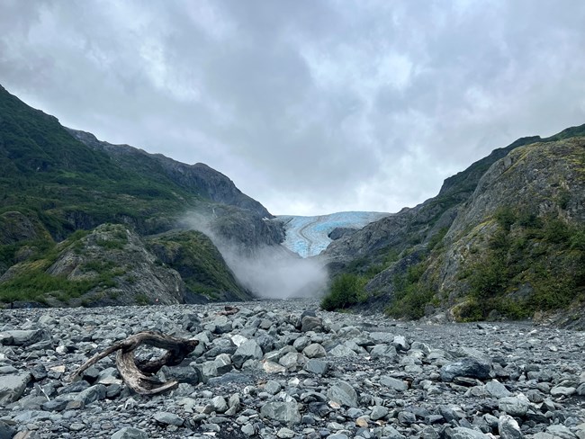 A foreground of rocks with Exit Glacier in the distance between green mountains.