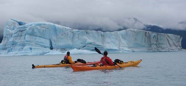 Two kayakers float in the water in front of a giant iceberg.