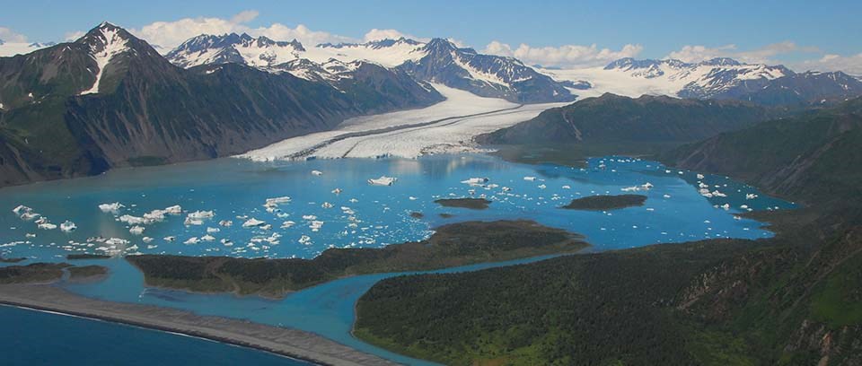 Large lagoon, surrounded by mountains, in front of Bear Glacier with white iceberg floating in the water.