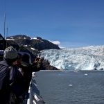 Two visitors take pictures of a tidewater glacier from the deck of a tour boat. Two visitors take pictures of a tidewater glacier from the deck of a tour boat.
