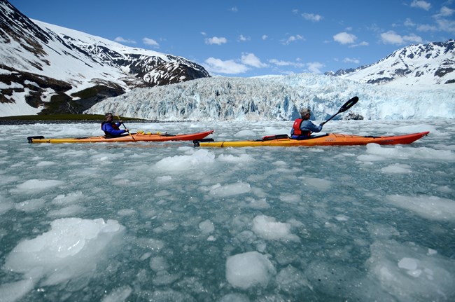 Two kayakers paddle in front of glacier