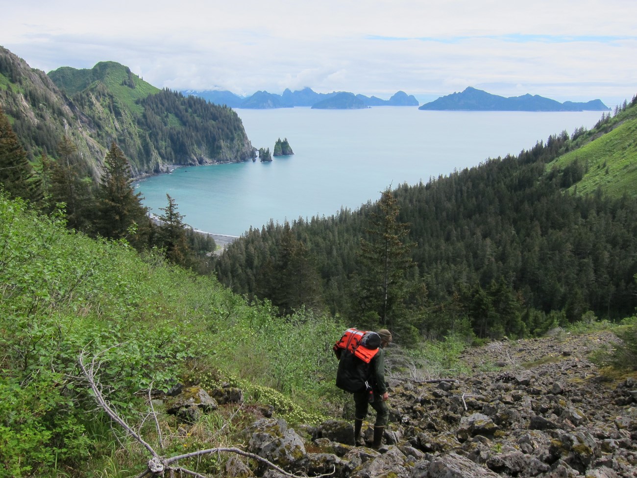 A hiker with large backpack steps down rocky terrain with a cove surrounded by steep mountains below.
