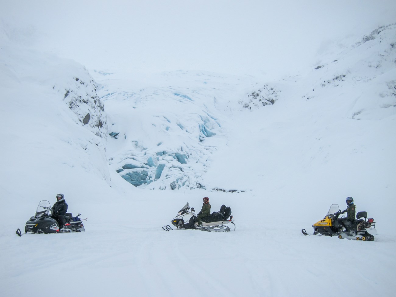 Three snowmobiles with riders wearing helmets in front of Exit Glacier under snow