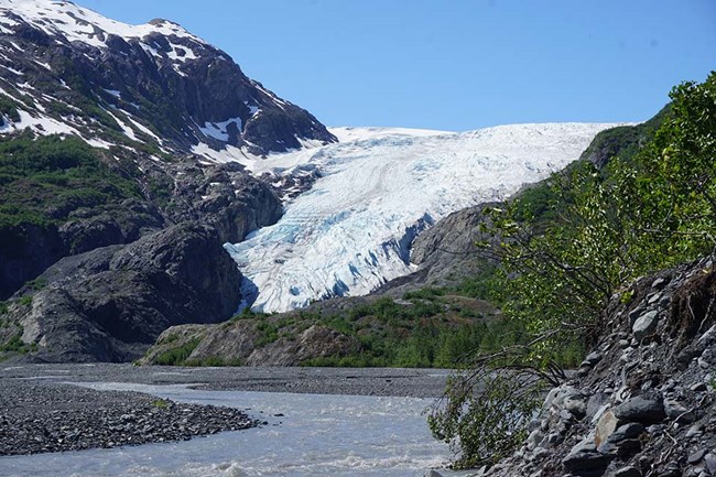 A glacier sits in a valley of grey rock, with a stream flowing into the foreground.