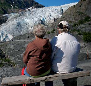 A couple sit on a bench located on the trail to the edge of Exit Glacier, and look to the glacier off in the distance. A couple sit on a bench located on the trail to the edge of Exit Glacier, and look to the glacier off in the distance.