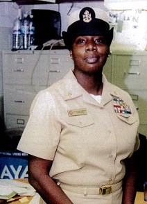 Black-skinned female stands in uniform in front of file cabinets inside. Wears a tan button down blouse with several colored tags pinned to chest. Wears fitted white hat with side brims that point up. Symbol of anchor on hat.