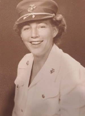 Portrait of white-skinned female in uniform. White blouse with gold buttons down front and on 2 front pockets. Fitted small round hat with front brim. Thin rope wraps around hat and dangles on brim. Matching insignia is on hat, and both collars of blouse.