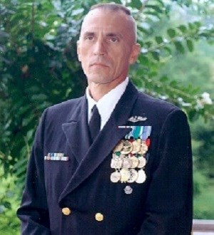 White-skinned Man stands tall in uniform outside in front of a tree. Wears blue formal jacket with shirt and tie underneath. 12 medals are pinned to his jacket in neat 3 x 4 rows.