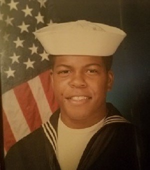 Portrait of black-skinned man in front of American flag. He wears a navy blue shirt with a collar that continues in back. He wears a white shirt underneath. A fitted white hat with full brim points up to sky.