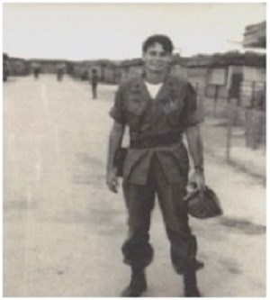 Man with dark hair and goatee stands on dirt street in uniform. He wears baggy pants tucked into boots. Long-sleeve, button-up shirt rolled up to his elbows with an ammunition belt around waist. He holds his hat.
