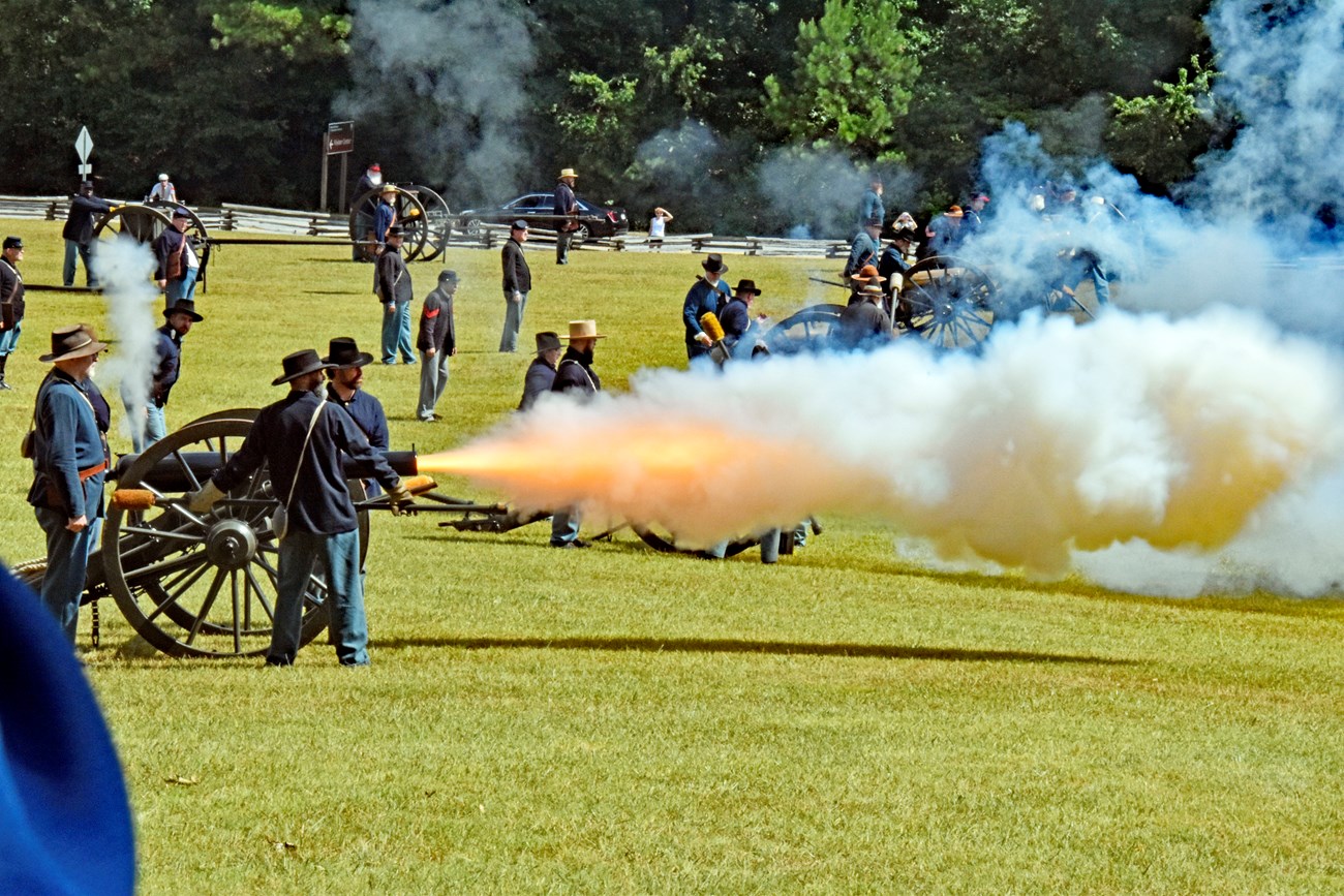 Group of people dressed in Civil War uniform stand in short grassy field firing cannons. Smoke and fire are coming out of cannons. Green trees and wooden fence seen in background.