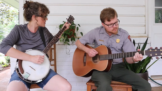 Person sits playing banjo while ranger sits playing guitar.