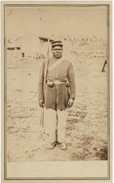 Black man stands at attention in military uniform in the dirt. He holds a musket and carries a satchel across his chest.