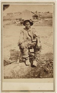 Black man sits on a chair outside on dirt. Wears a brimmed hat, torn long-sleeved shirt, and boots