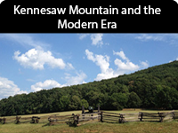 Densely tree-covered mountain sits under a blue sky with a few clouds and behind a picket fence.