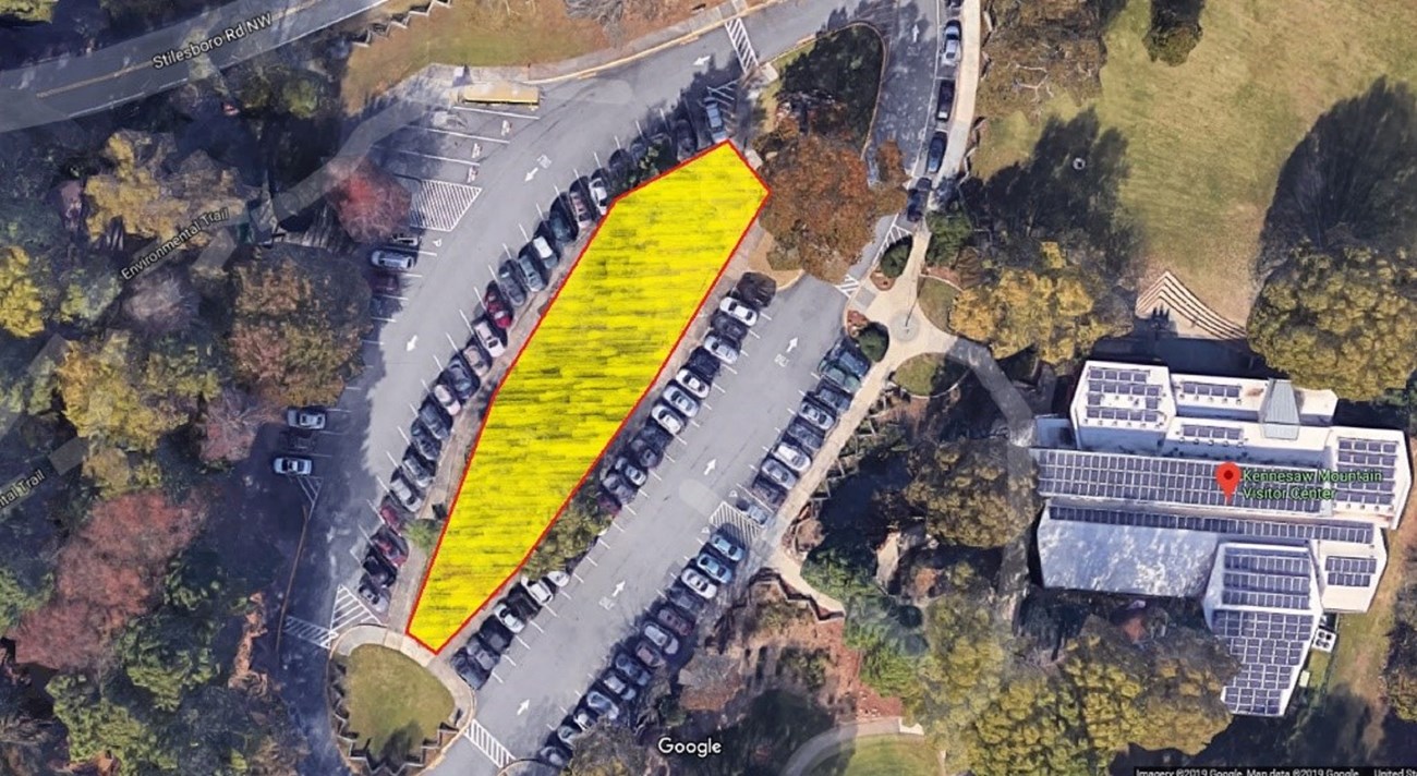 Yellow highlights fill the grass island between the center rows of cars in the parking lot east of rectangular building. Parking lot and building are connected by a sidewalk on the northwest side of lot. Grass and trees line each side of sidewalk.