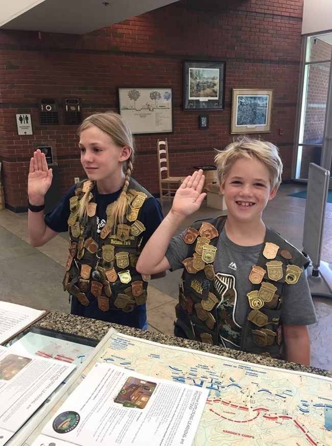 Two children with many Jr Ranger badges raise their right hands at a desk with park maps on it.