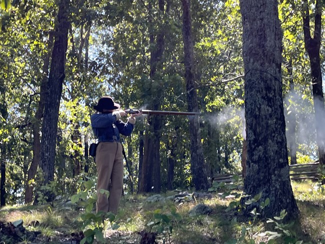 Living history demonstration of Civil War soldier firing rifle. Person stands surrounded by trees while fiiring gun which emits smoke.