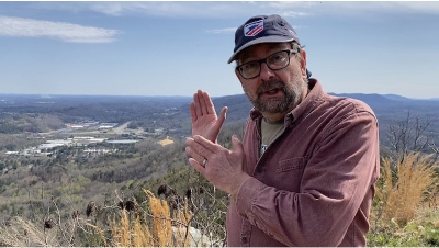 Man stands outdoors jesturing to far off town with hands