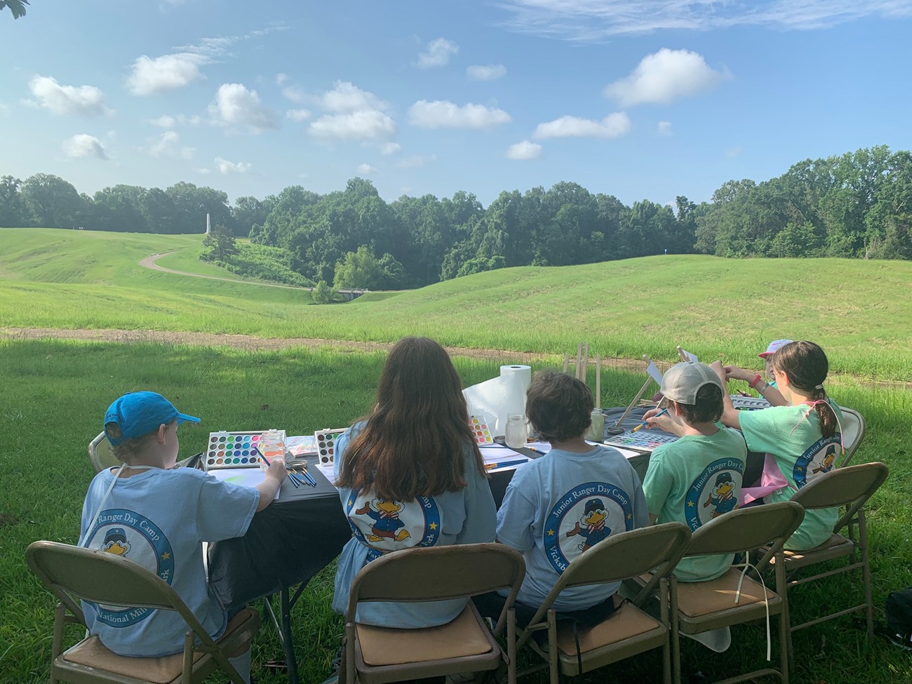 Five children sit at a table outdoors painting. Green field with tress and bright blue skies with white clouds seen in distance.