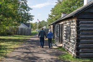 A couple holding hands walks down a path next to a historic wooden structure.