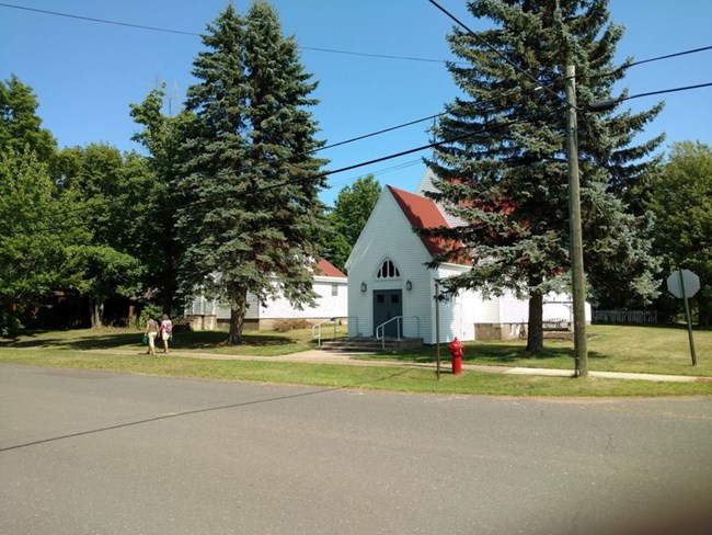 Two people walk down a sidewalk in front of a white-sided, red-roofed church.