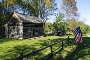 Restored log dwellings are surrounded by green grass and a fence with an American flag attached to it.