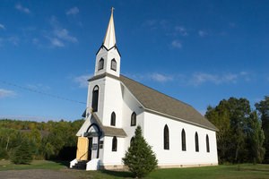 A white exterior church with a narrow steeple surrounded by forest.