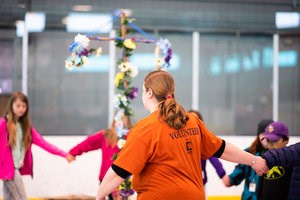 Adults and students holding hands dance around a pole with flowers on it.