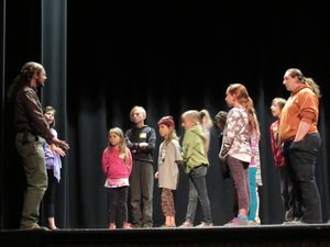 A group of students listen to directions from an adult on a stage.
