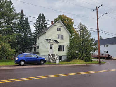 A two-story front-gabled frame house has a car parked alongside it. Stairs reach the elevated front door.