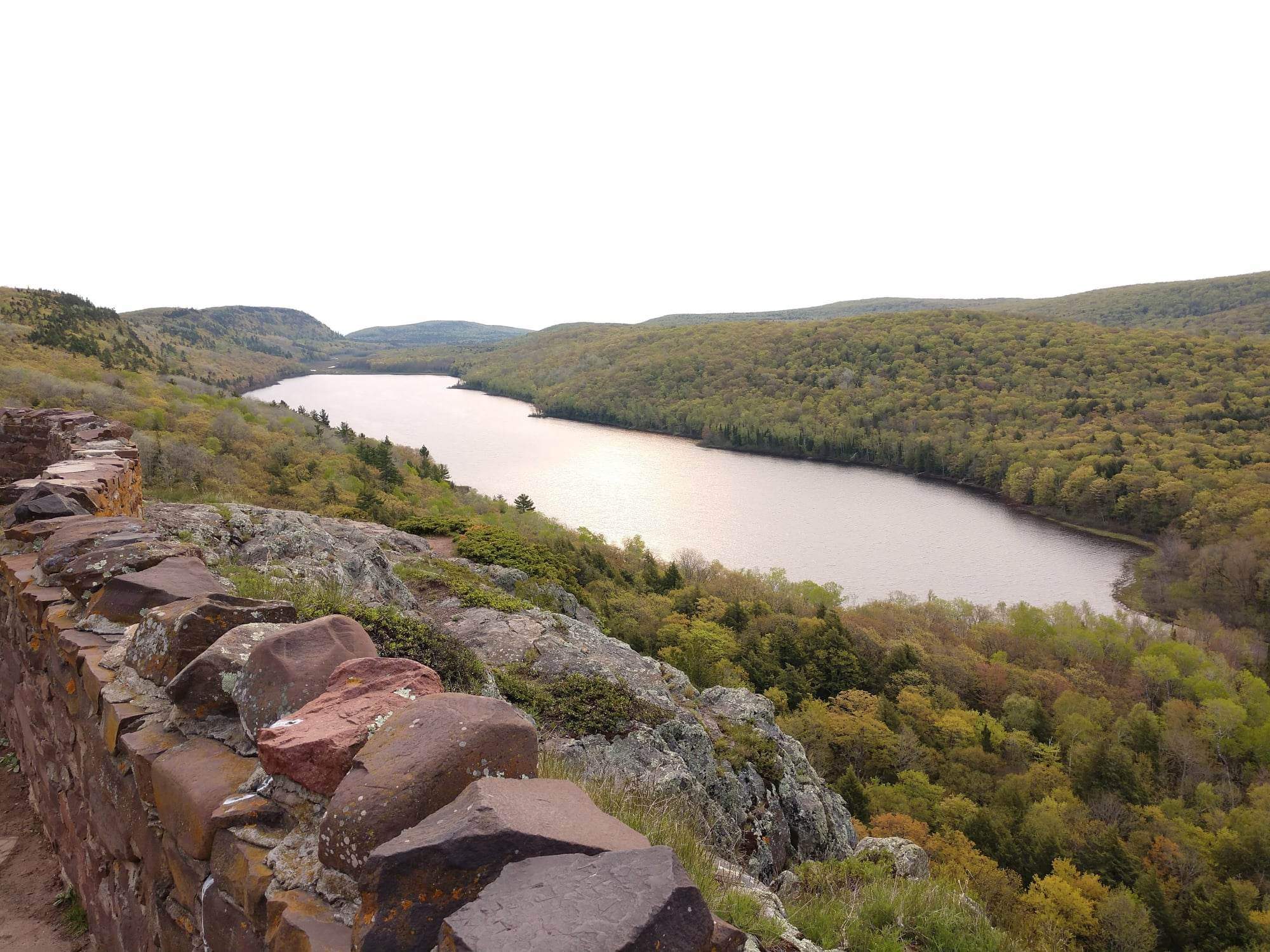 A landscape featuring a large lake on a cloudy day surrounded by rolling hills of dense forest and rocky outcroppings.